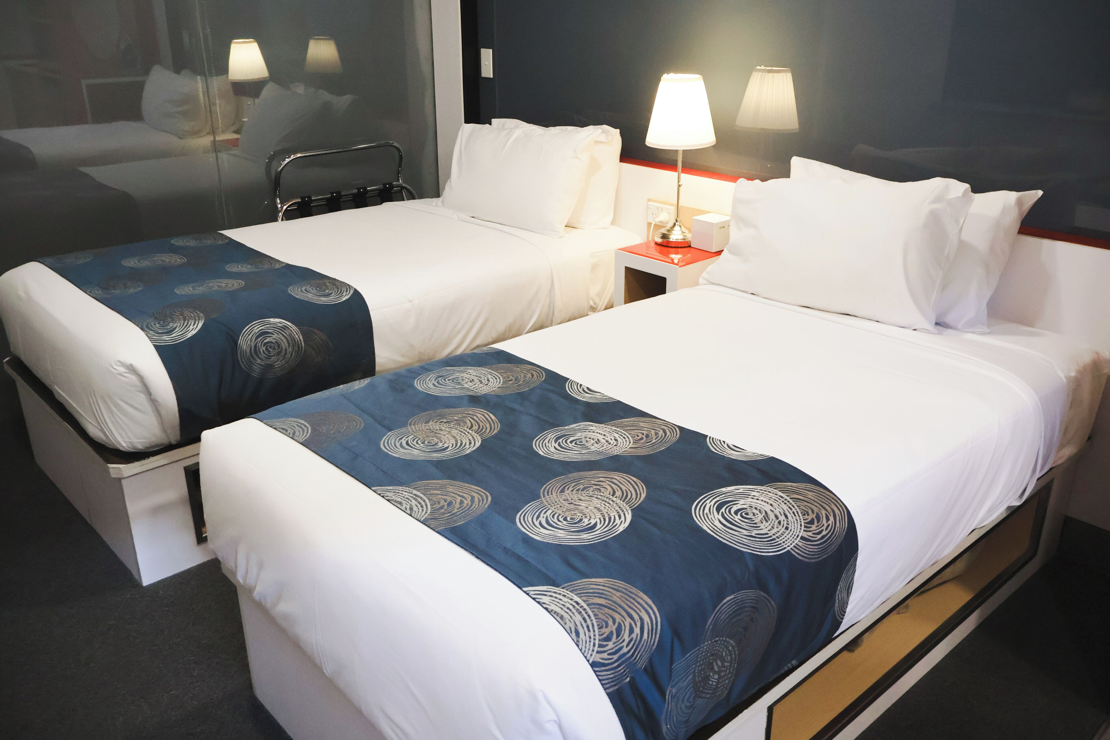 A hotel room with two single beds, each with a blue patterned bedspread and white pillows. There is a nightstand with a lamp between the beds.