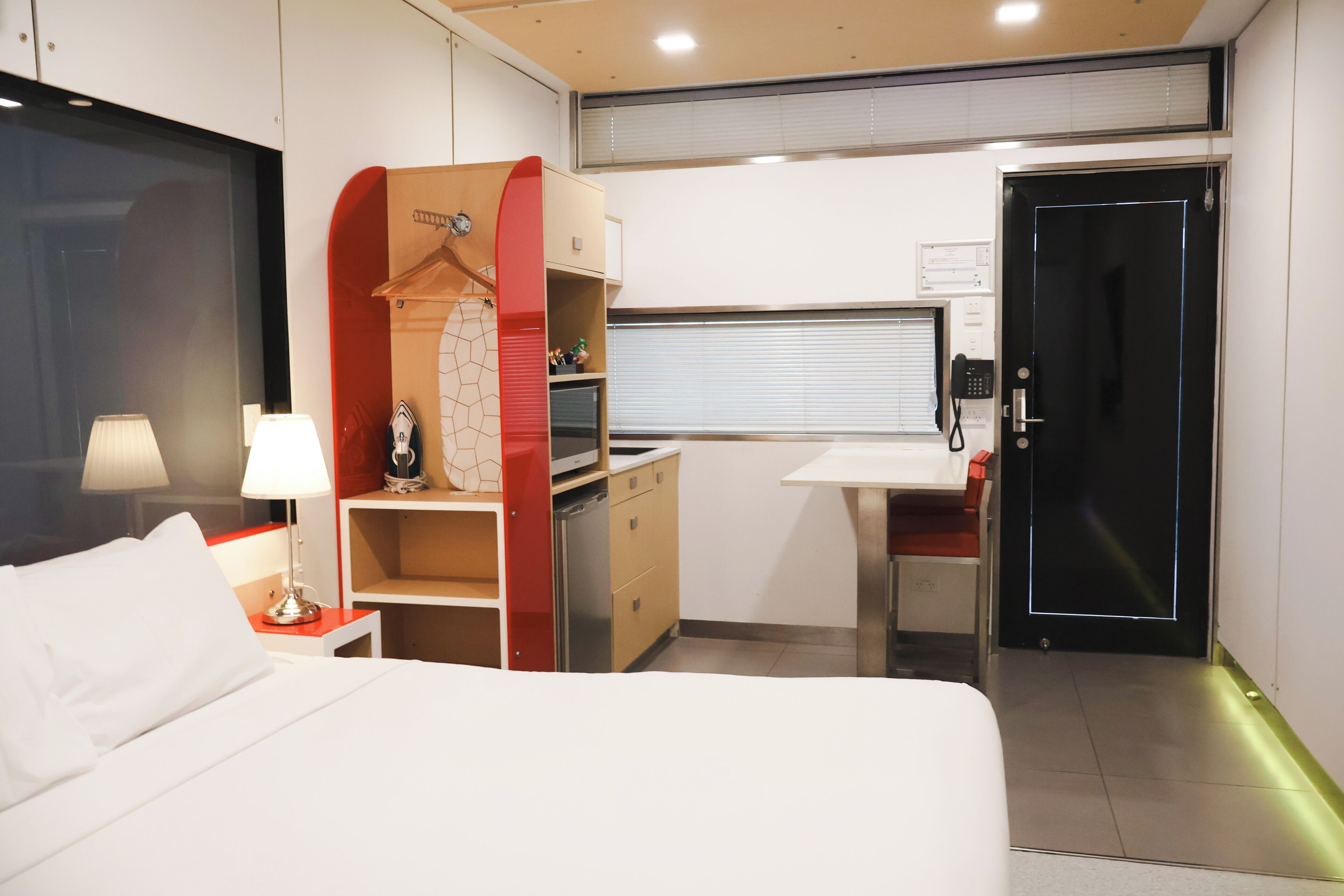 Una habitación de hotel con cama, escritorio, armario, lámpara y puerta.