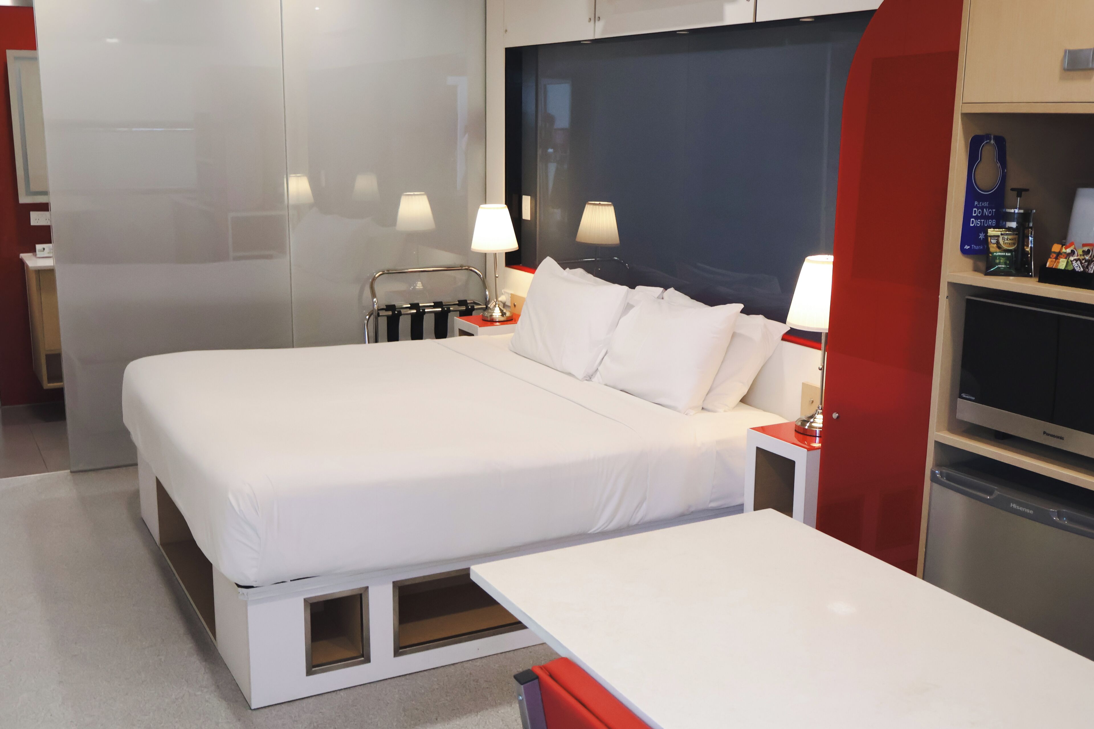 A modern hotel room with a large bed, bedside tables, a nightstand, a TV, and a minibar.