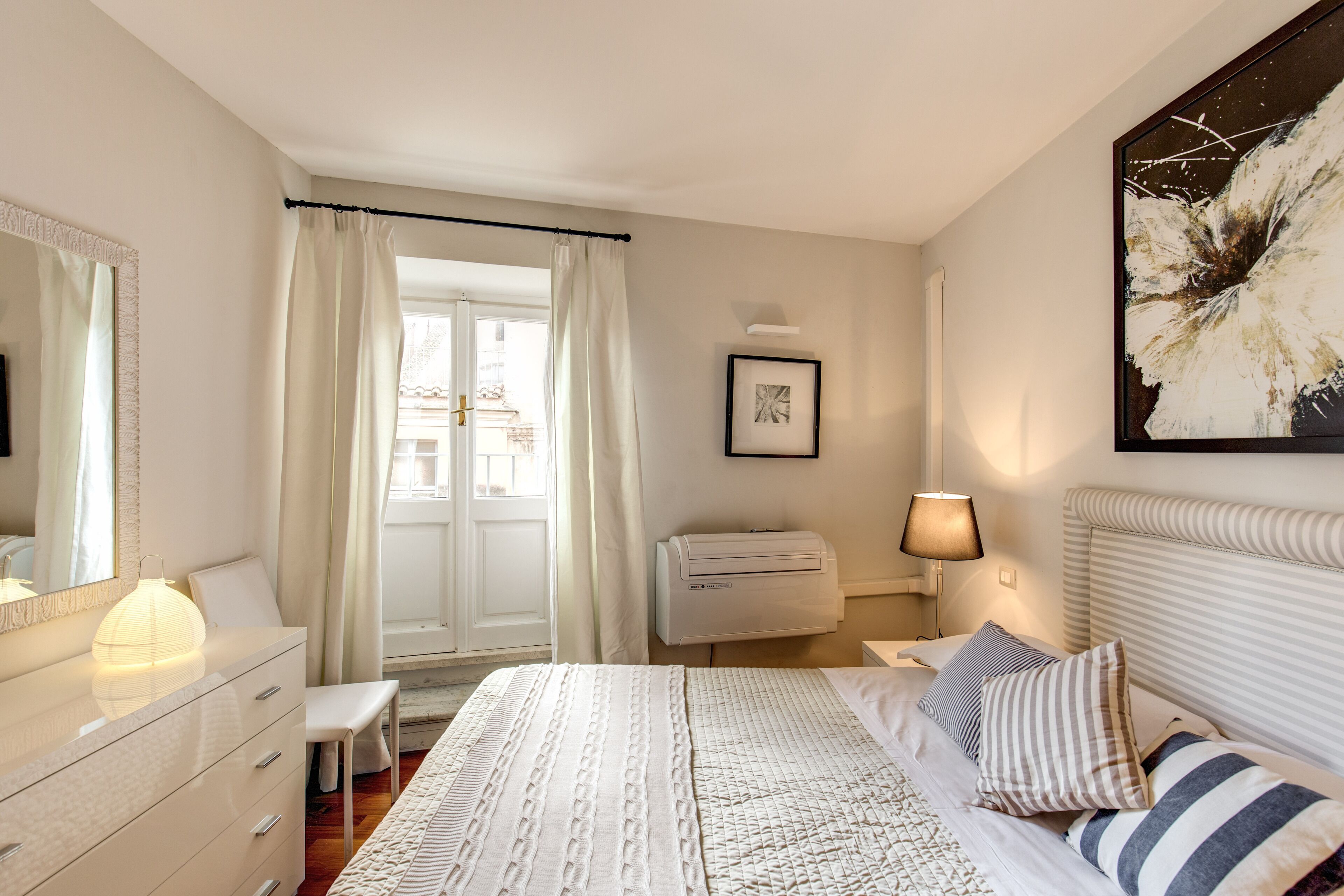 A bedroom with a bed, bedside table, lamp, air conditioning unit, and a window with curtains.