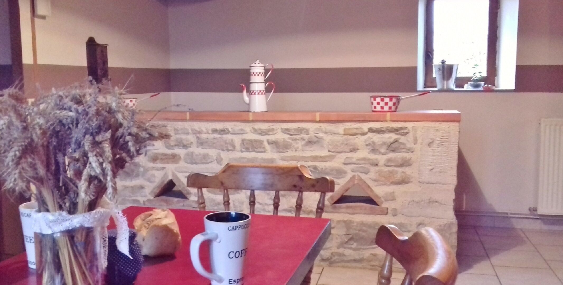Una chimenea de piedra con una bandeja blanca y roja a cuadros encima, un banco de madera y una mesa con mantel rojo, un jarrón de flores secas y dos tazas de café.