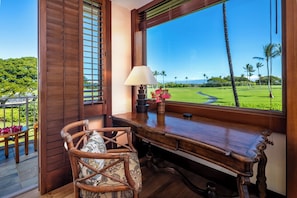 A wooden desk with a view of a golf course, a wicker chair, and a lamp.