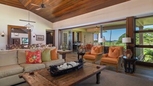A spacious living area with a wooden ceiling, a sofa with colorful cushions, a coffee table, and a view through large windows. - Fairway Villa 120A: Charming w/Shared Pool, AC, 2 Golf Carts & Lanai Ocean Views (Kailua Kona)