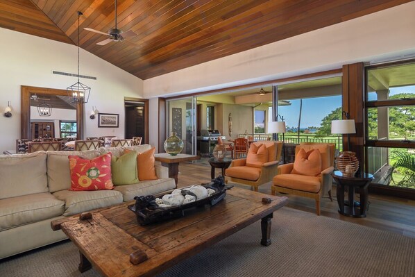 A spacious living area with a wooden ceiling, a sofa with colorful cushions, a coffee table, and a view through large windows. - Fairway Villa 120A: Charming w/Shared Pool, AC, 2 Golf Carts & Lanai Ocean Views (Kailua Kona)
