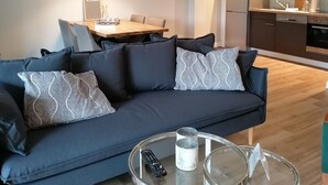 A modern living room with a dark blue sofa, a glass coffee table, and a dining area with a wooden table and chairs. - Apartment with hotel comfort (Bad Rothenfelde)