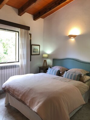 Un dormitorio con una cama grande, una mesita de noche con lámpara, un cuadro enmarcado en la pared y una ventana con cortinas.