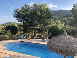 Piscina con una pérgola y asientos al aire libre, rodeada de árboles y montañas al fondo.
