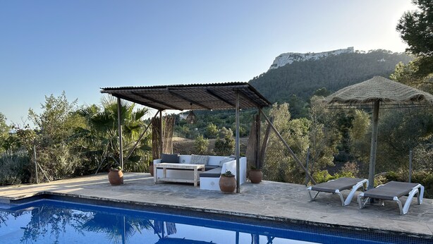 Un área de piscina con zona de estar cubierta, sillas de descanso y vistas a un paisaje montañoso.