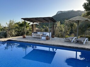 Un espace piscine doté d’un coin salon couvert, de chaises longues et offrant une vue sur un paysage montagneux.