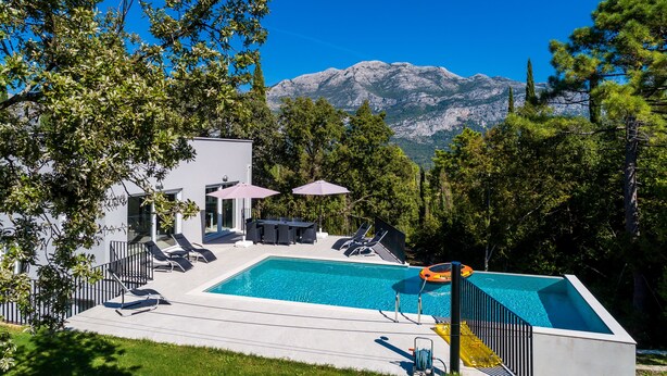 Un espace piscine moderne, doté de chaises longues et de parasols, entouré d’arbres et avec des montagnes en toile de fond.