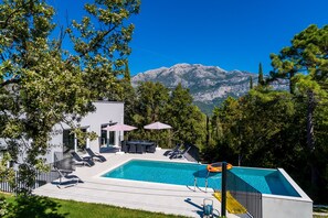 A modern pool area with lounge chairs and umbrellas, surrounded by trees and mountains in the background.
