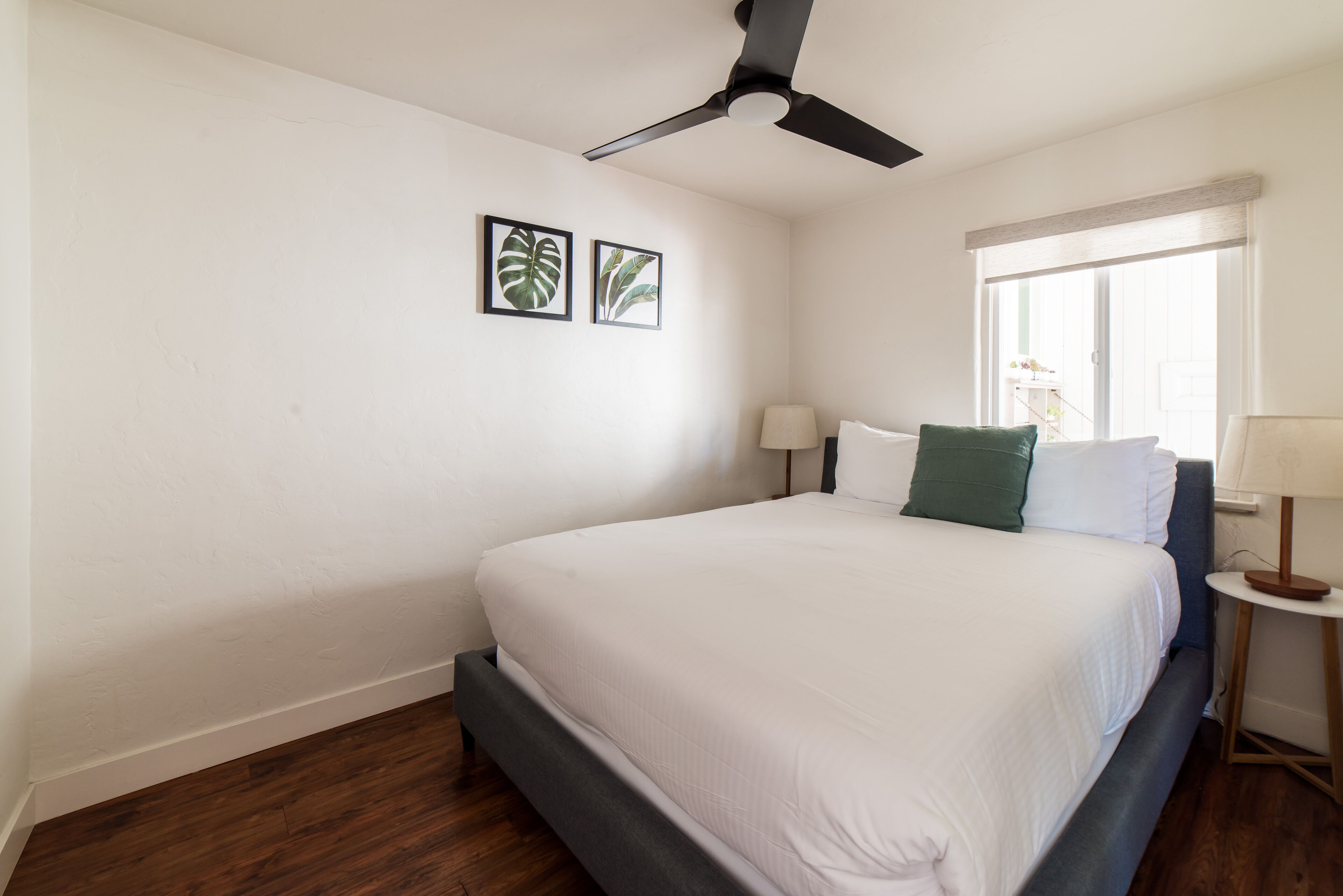 Une chambre à coucher avec un lit, un ventilateur de plafond, deux tableaux encadrés, une fenêtre donnant sur un paysage agréable et une table de chevet avec une lampe.