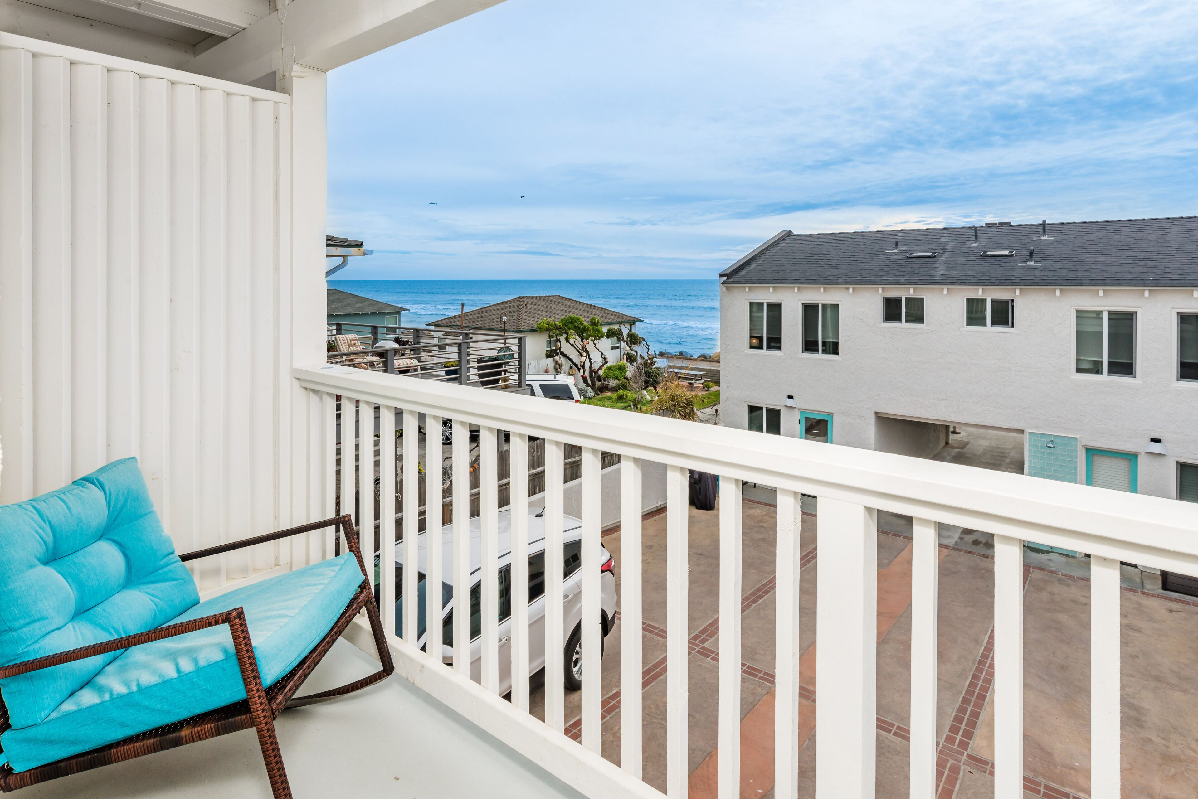 Un balcon avec une balustrade blanche, une chaise bleue avec un coussin et une vue sur l’océan et les bâtiments.