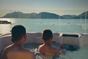 Zwei Personen in einer Badewanne mit Blick auf ruhiges Gewässer und ferne Berge.