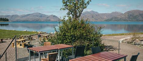 Zona de estar al aire libre con mesas y sillas, con vista a un lago tranquilo y montañas.