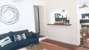Ein modernes Wohnzimmer mit einem dunkelblauen Sofa, einem Holztisch und einem weißen Teppich. Im Hintergrund ist eine Küche mit einer Hängelampe zu sehen.