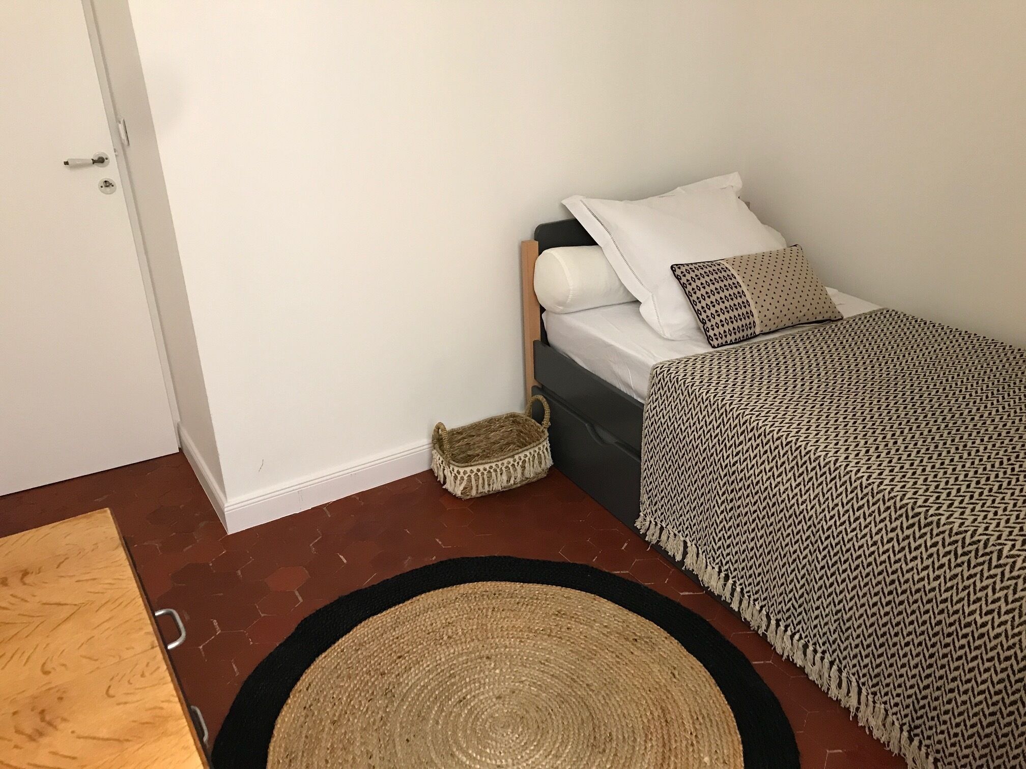 A bedroom with a bed, a bedside table, a small woven basket, and a circular woven rug.