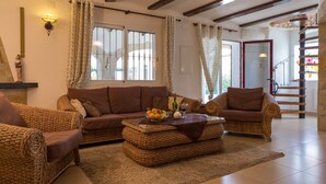 Uma sala de estar acolhedora com móveis de vime, uma mesa de centro com frutas e uma garrafa, e uma escada que leva a um andar superior.