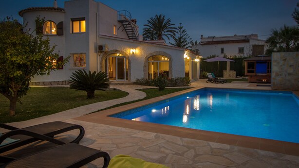 Piscina com espreguiçadeiras e área de pátio, cercada por um jardim ajardinado e um edifício com janelas em arco e iluminação externa.
