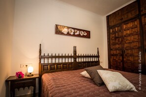 Een slaapkamer met een houten hoofdbord, een nachtkastje met een lamp, een bloemverf schilderij aan de muur, en een grote houten deur.