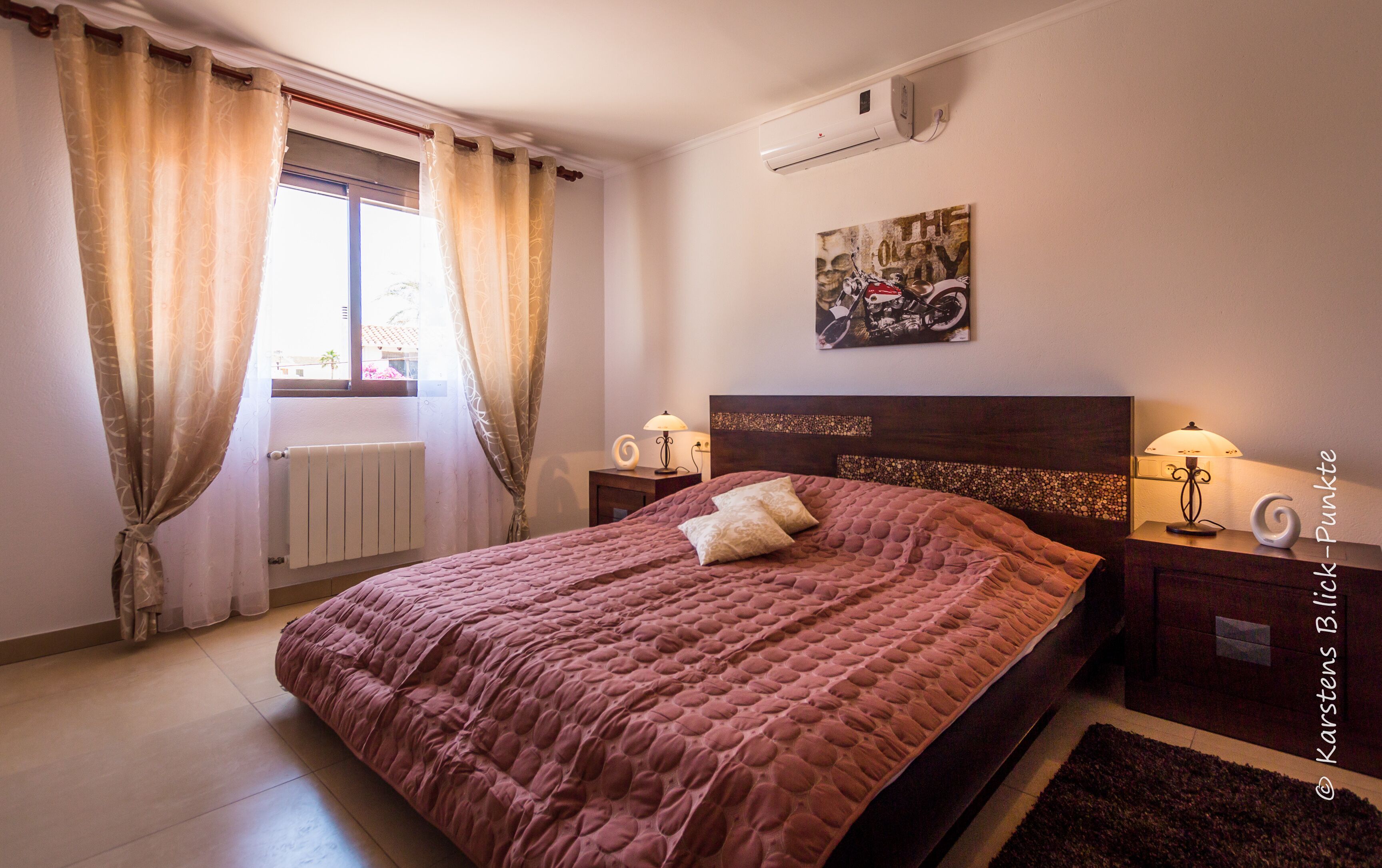 Un dormitorio con cama, dos mesitas de noche, un radiador, una ventana con cortinas, un aire acondicionado y una obra de arte colgada en la pared.