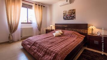 Un dormitorio con cama, dos mesitas de noche, un radiador, una ventana con cortinas, un aire acondicionado y una obra de arte colgada en la pared.