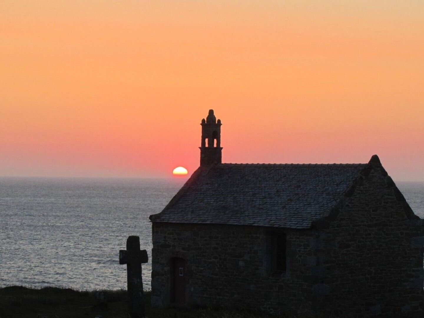 A stone building with a cross, a bell tower, and a sunset over the sea.