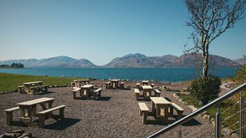 A lakeside picnic area with wooden tables and benches, a gravel path, and a view of mountains in the distance.
