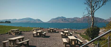 A lakeside picnic area with wooden tables and benches, a gravel path, and a view of mountains in the distance.