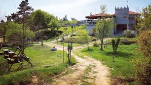 Un edificio rústico con techo de tejas rojas, rodeado de un césped con un sendero de tierra que lleva a una mesa de picnic y un columpio.