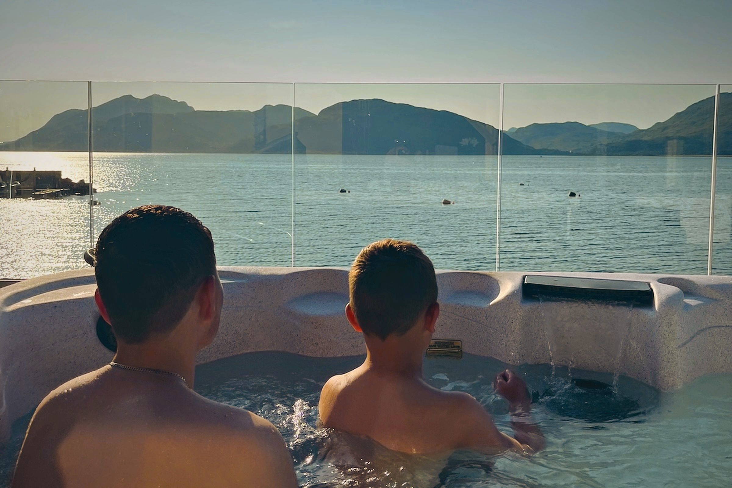 Two individuals in a hot tub with a view of a calm body of water and distant mountains.