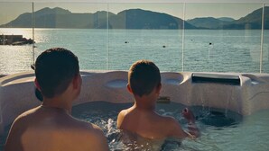 Dos personas en un jacuzzi con vista a un cuerpo de agua tranquilo y montañas lejanas.