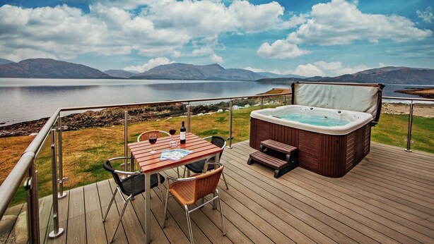 Una terraza con jacuzzi, mesa y sillas con vista a un lago y montañas al fondo.