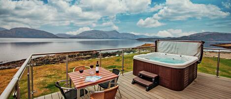 Una terraza con jacuzzi, mesa y sillas con vista a un lago y montañas a lo lejos.