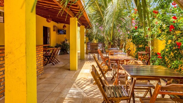 An outdoor patio with wooden tables and chairs, yellow walls, and a palm tree. - Pousada Amarante praia (São Miguel do Gostoso)