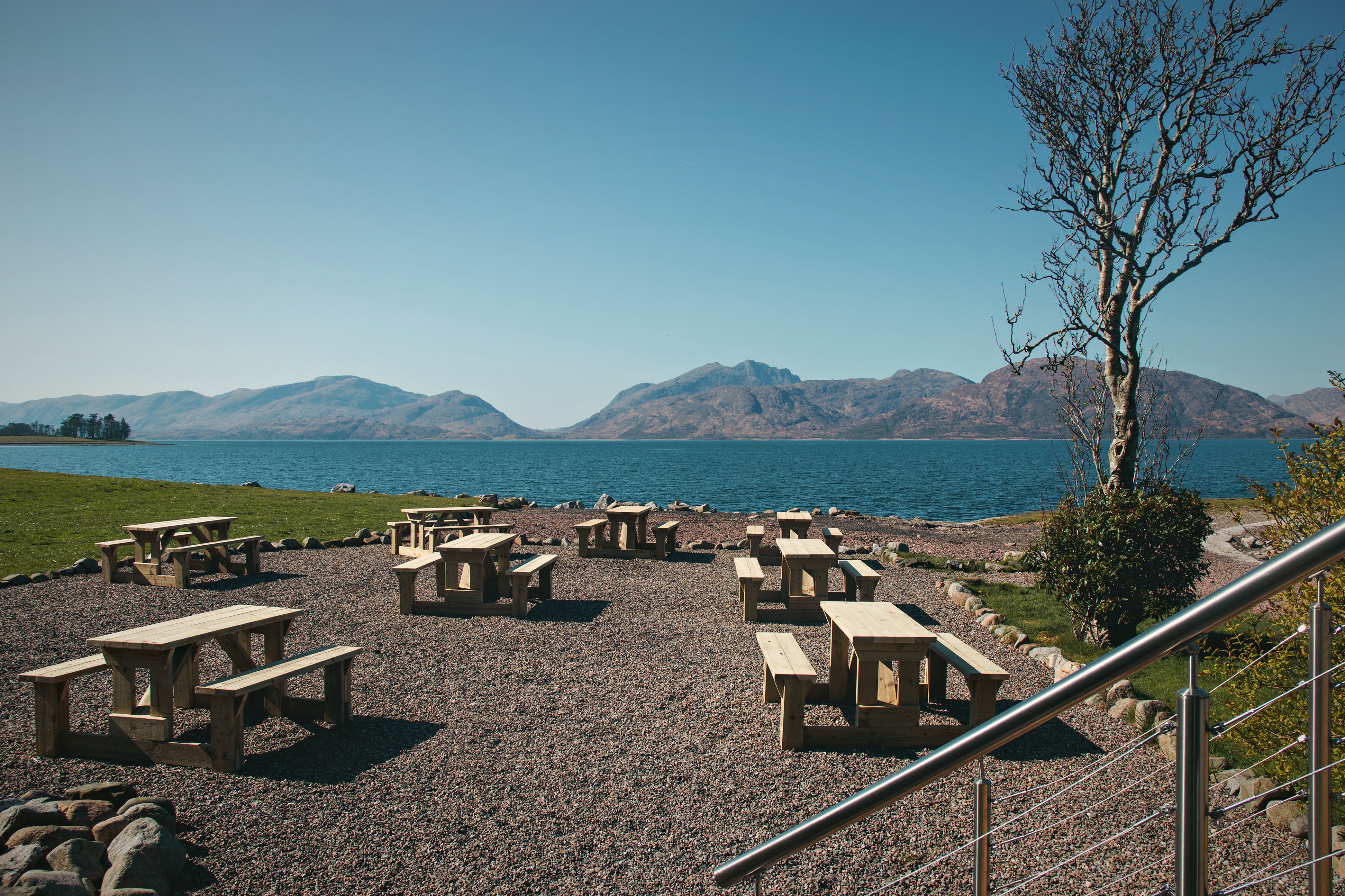 Un área de picnic junto al lago con mesas y bancos de madera, un sendero de grava y vistas a montañas distantes.