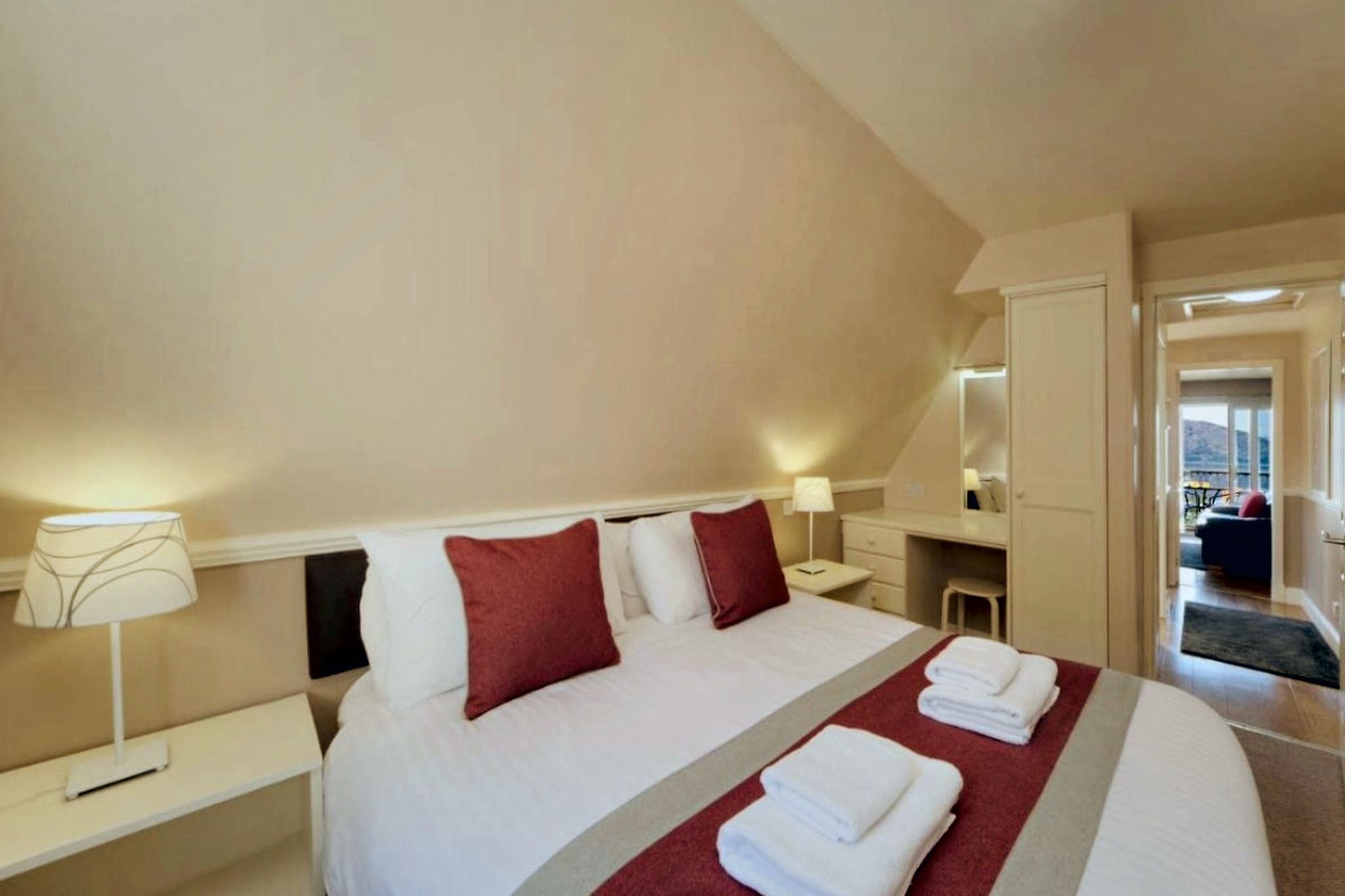 A hotel room with a large bed, two bedside tables, a nightstand, a desk, and a chair. There is a window with a view of the outside.