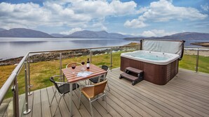 A deck with a hot tub, table, and chairs overlooking a lake with mountains in the background.