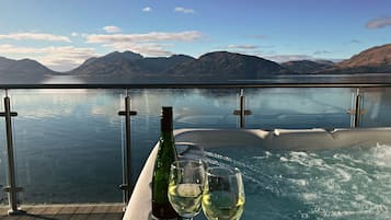 Un bain à remous, une bouteille de vin et deux verres sur une terrasse en bois donnant sur un lac paisible, avec des montagnes en arrière-plan.