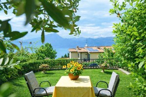 A garden with a table and chairs, a flower pot, and a view of a house with solar panels and mountains in the background. - B&B Musik Suite with a View of Lake Garda (Torri del Benaco)