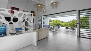 Een moderne receptiebalie met een wit bureau, een decoratieve wand en uitzicht op het buitenterrein met zitplaatsen.