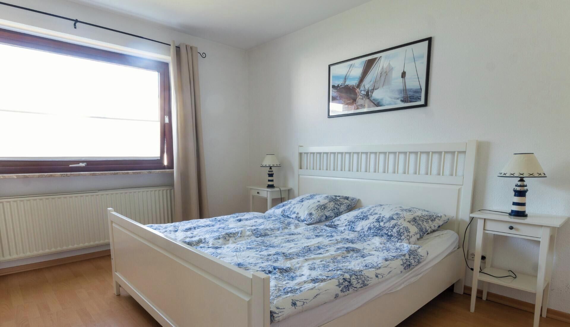 A bedroom with a bed, two bedside tables, a window with curtains, a radiator, and a painting on the wall.