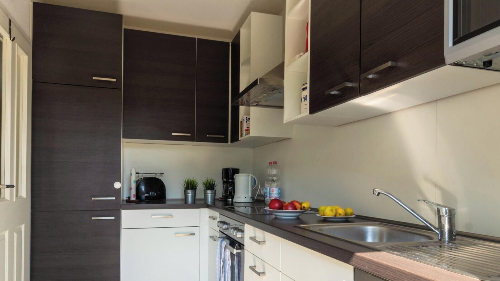 Una cocina compacta con armarios oscuros, un lavamanos de acero inoxidable y una encimera con hervidor, cafetera y fruta.
