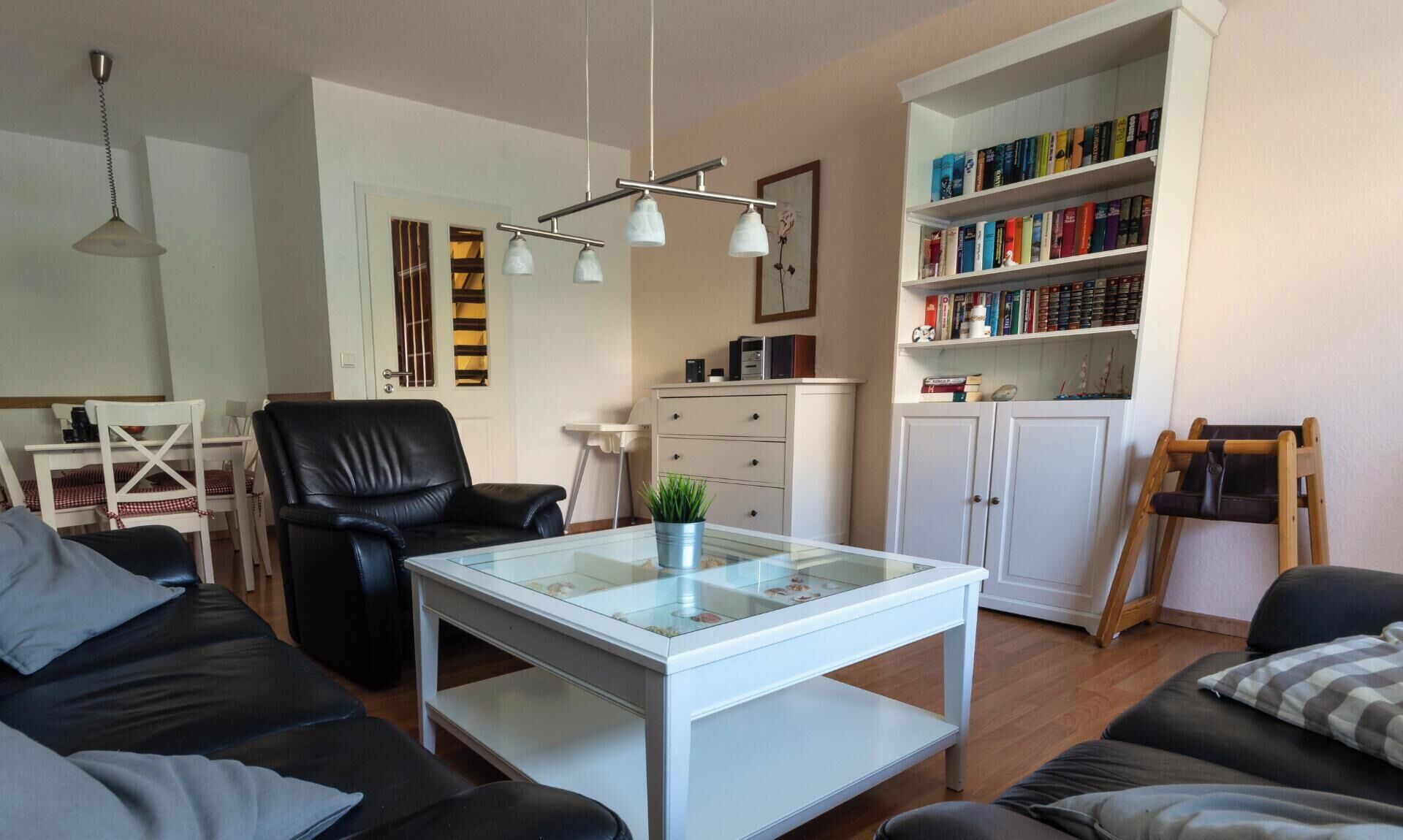 A living room with a glass-top coffee table, a black leather sofa, a white cabinet with books, and a wooden chair.