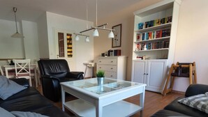 A living room with a glass-top coffee table, a black leather sofa, a white cabinet with books, and a wooden chair. - 🌊 Holiday home for 6 in Grömitz | 2 bathrooms, garden & beach chair on the beach (Grömitz)
