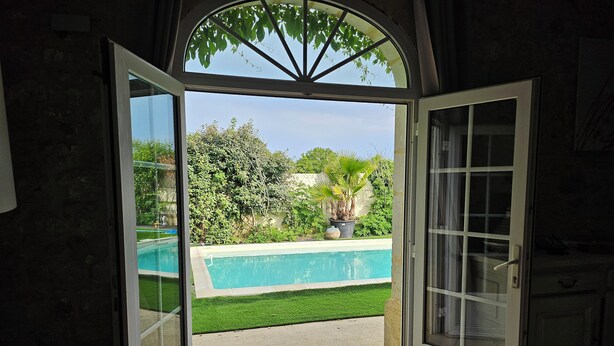 Una puerta abierta que da a un patio trasero con piscina, rodeado de vegetación y un cielo despejado.