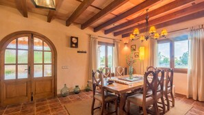 A dining room with a wooden table, chairs, and a chandelier. There are windows with curtains and a view of greenery outside. - Sa Tanqueta Rustica, Country House 5StarsHome Mallorca (Inca)
