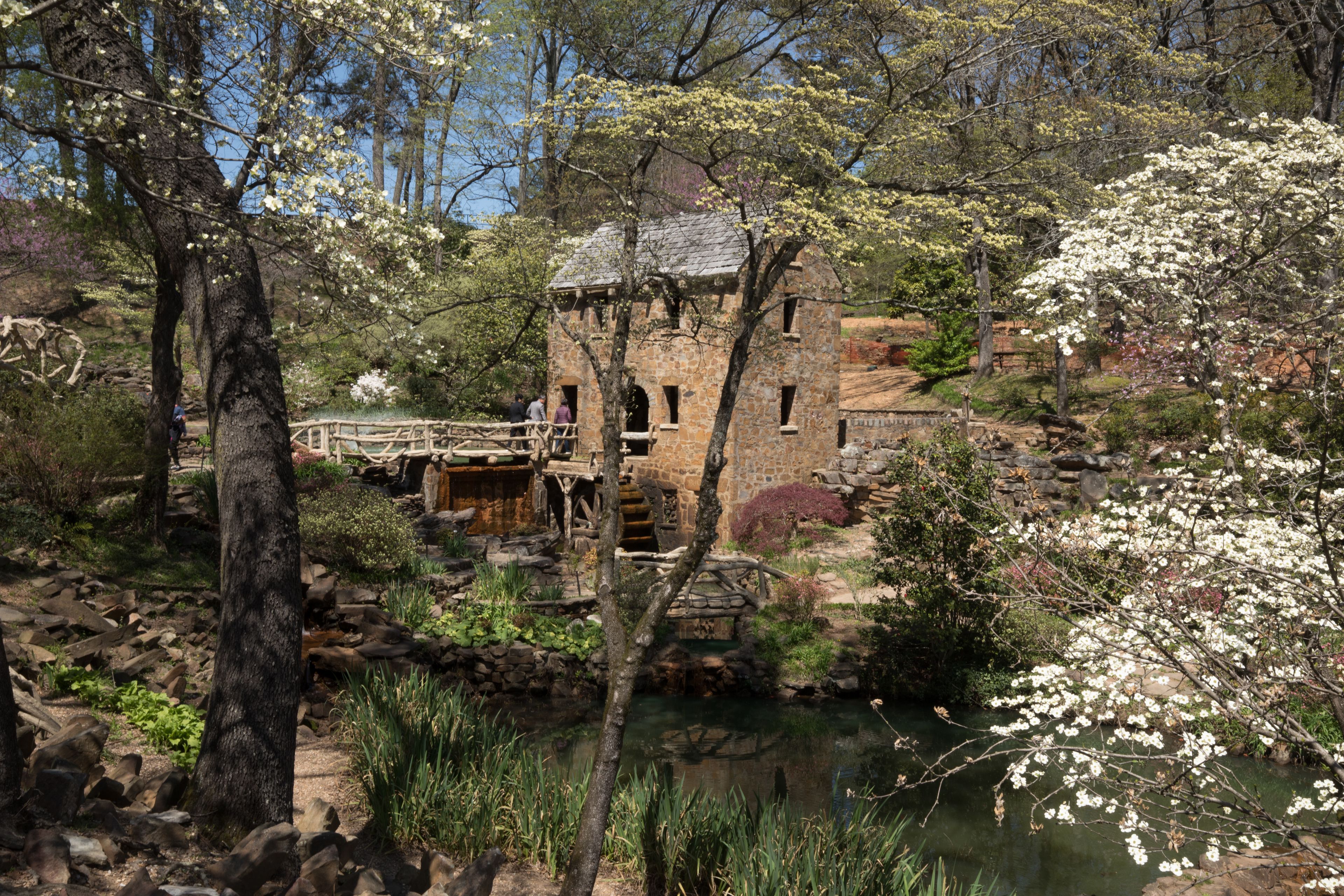 A stone building with a wooden bridge over a pond, surrounded by trees and vegetation.
