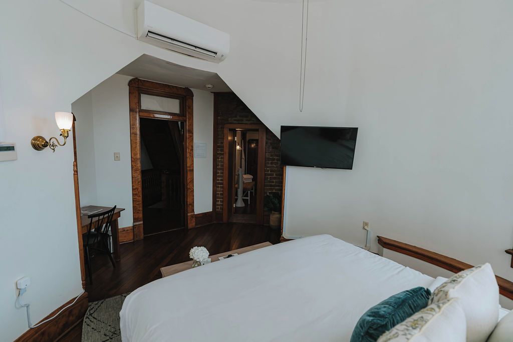 Quarto com cama, televisão na parede, ar condicionado, escrivaninha com cadeira e uma mesinha com um vaso.