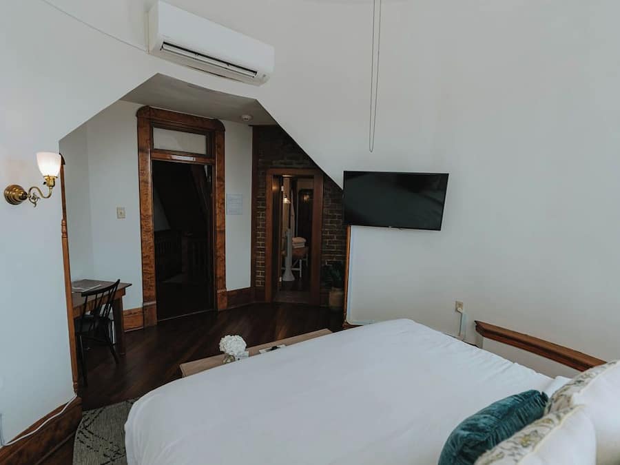 Quarto com cama, televisão na parede, ar condicionado, escrivaninha com cadeira e uma mesinha com um vaso.